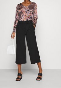 Langärmliges, V-Ausschnitt-Oberteil mit einem bunten Paisleymuster, kombiniert mit hoch taillierten, weiten schwarzen Hosen und schwarzen Slipper-Heels, hält eine weiße Handtasche.
