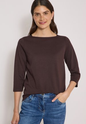 Femme aux longs cheveux foncés portant un haut brun foncé à manches trois-quarts et un jean bleu, une main dans la poche, souriant à la caméra.