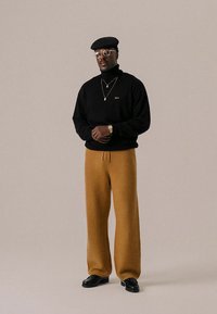 Black knit sweater, brown textured wide-leg pants, black shoes, round glasses, and a black cap with a gold detail. Minimalist jewelry accents.