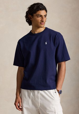 Νέος άνδρας με σγουρά σκούρα μαλλιά, που φοράει μπλε κοντομάνικο πουκάμισο και λευκό παντελόνι, κοιτάζει ελαφρώς πλάγια.