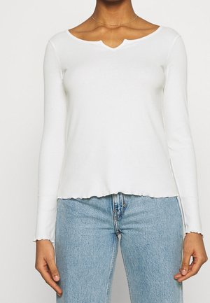 Person trägt ein weißes, langärmliges, geripptes Oberteil mit wellenförmigem Saum und Ausschnitt, kombiniert mit hellblauen Denim-Jeans.