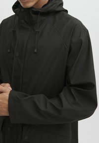Juodas vandeniui atsparus džemperis su kapišonu, turintis reguliuojamus sujungimus, užsegimus ant spaudžių ir lygią tekstūrą. Sukurtas su raglano rankovėmis.