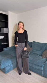 Haut à manches longues noir, pantalons à jambes larges gris, sac à main noir, debout sur un canapé texturé bleu dans une pièce lumineuse avec des étagères en arrière-plan.