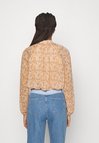 Blusa floral de Manga Larga en naranja claro y crema, con pliegues en la parte trasera y un dobladillo con forma de ondas. Combinada con unos jeans azul de talle alto.