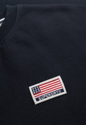 Un sweat-shirt bleu marine en tissu avec un col côtelé. Il présente une étiquette rectangulaire blanche avec un drapeau des États-Unis et "SUPERDRY" en texte bleu.