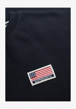 Sudadera de color marino con un cuello acanalado. Presenta un parche rectangular blanco con una bandera de EE. UU. y "SUPERDRY" en texto azul.