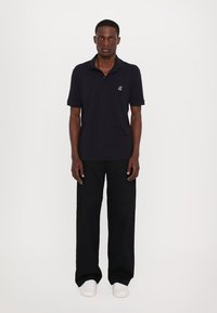 Svart polo skjorte med korte ermer, med en liten logo på brystet; kombinert med vide svarte bukser og hvite sneakers.