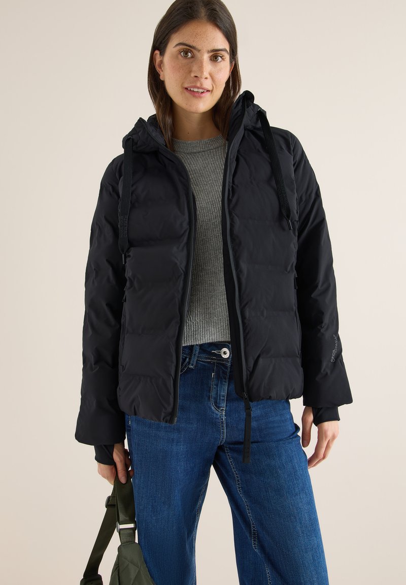 Sort pufferjakke med hætte, med en frontlynlås og justerbare snore. Sammen med en grå sweater og blå jeans.