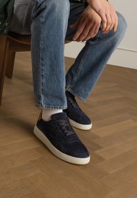 Sneakers in camoscio navy con suola in gomma bianca e accenti del tallone in beige a contrasto, abbinate a jeans di denim azzurro chiaro e calze bianche.