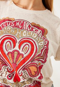 Mujer vistiendo una camiseta beige con un diseño de corazón colorido y texto en tonos rojo, naranja y blanco.