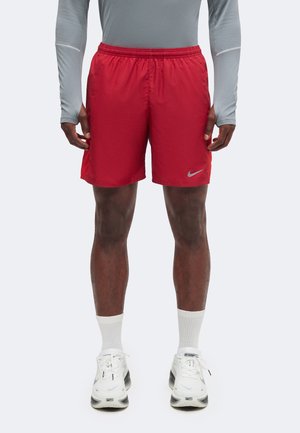 Mand iført lyse røde Nike atletikshorts, hvide sokker og hvide Nike sneakers står mod en ensfarvet baggrund.