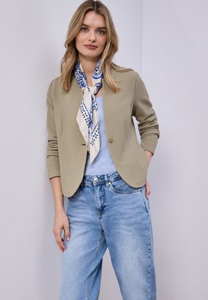 Femme portant un blazer texturé beige, un haut bleu clair, un foulard à motifs bleus autour du cou, et un jean bleu clair, debout avec une main dans la poche.