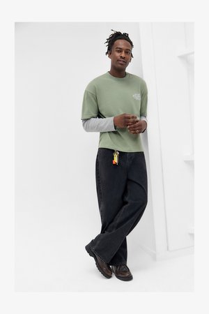 Man with dreadlocks leaning against white wall, wearing layered green and gray shirts, black wide-leg jeans with keychain, and brown shoes.