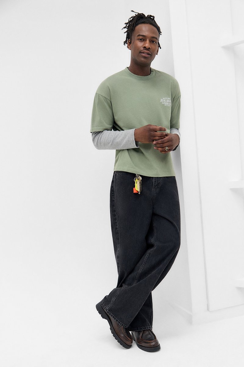 Mann mit Dreadlocks, der an einer weißen Wand lehnt, trägt geschichtete grüne und graue Shirts, schwarze weite Jeans mit einem Schlüsselbund und braune Schuhe.