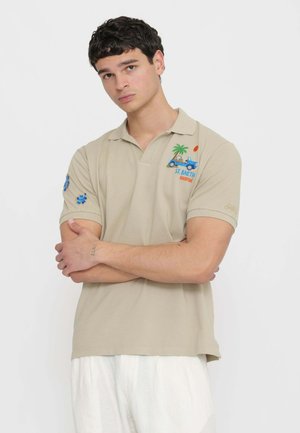 Giovane uomo con polo beige con macchina blu ricamata, palma, sole e testo, in piedi con le braccia incrociate su sfondo bianco.
