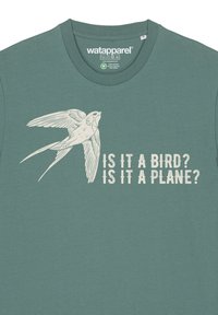 Grünes Baumwoll-T-Shirt mit einem weißen Schwalbenmotiv und dem Aufdruck "IST ES EIN VOGEL? IST ES EIN FLUGZEUG?" darunter in fetter Schrift.