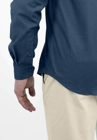 Marineblaues Langarmhemd mit Knopfmanschette, das eine dezente Textur aufweist. Kombiniert mit hellbeigen Hosen, die einen abgerundeten Saum haben.