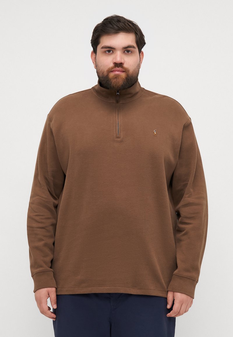 Bruine fleece pullover met een hoge kraag en een ritssluiting tot de helft. Heeft lange mouwen en een klein logo op de borst. Zachte textuur.