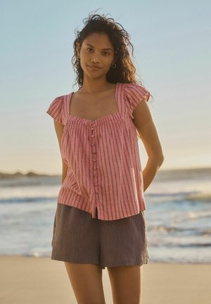 Frau mit langen lockigen Haaren, die ein pink gestreiftes, geknöpftes Oberteil und dunkle Shorts trägt, steht an einem Strand nahe ruhiger Meereswellen.
