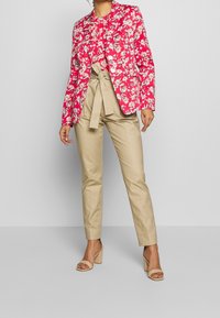 Blazer con motivo floreale in un vivace rosa, abbinato a pantaloni beige a vita alta con cintura annodata. Styling con sandali con tacco nude.