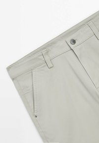 Lichtgrijze katoenen broek met een geplooide voorkant, twee voorzakken en een sluiting met een knoop aan de voorkant. Soepele textuur met subtiele stikseldetails.