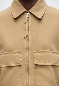 Κοντινό πλάνο μπεζ ζακέτας με φερμουάρ, με γιακά και δύο μεγάλα τσεπάκια με καπάκι στο στήθος, που φοριέται από άτομο με σκουρόχρωμο δέρμα.