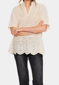 Blouse couleur crème avec un motif en œillets, décolleté en V, manches courtes à volants et ourlet festonné. Associée à un pantalon en denim foncé.