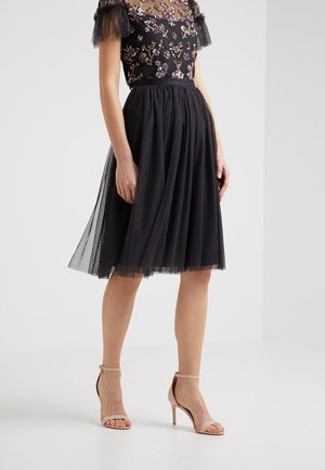 Femme portant un haut noir brodé de fleurs et une jupe en tulle noire, assortis à des sandales beige à talons hauts avec bride à la cheville.