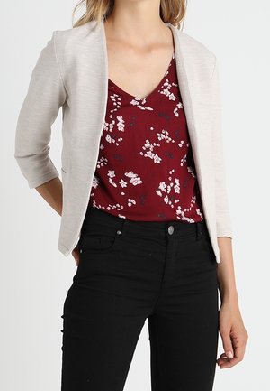 Femme portant une veste ouverte beige clair, un top à col en V floral bordeaux, et un pantalon noir ajusté avec une fermeture à bouton.