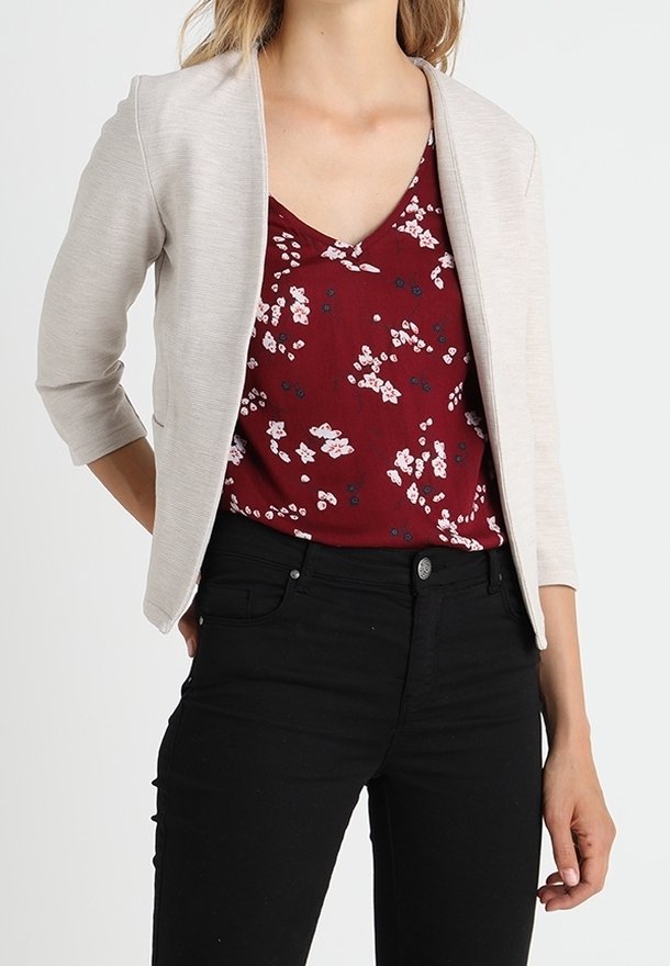 Femme portant une veste ouverte beige clair, un top à col en V floral bordeaux, et un pantalon noir ajusté avec une fermeture à bouton.