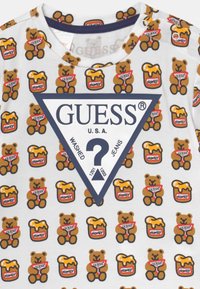 Witte korte mouwen shirt met een herhaald patroon van teddyberen en honinglegels. Centraal een marineblauwe driehoek met "GUESS®" prominent weergegeven.