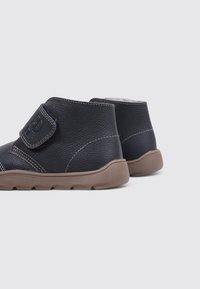 Unos botines negros de cuero para niños, con suelas de goma marrón y correas de velcro, sobre un fondo gris claro.