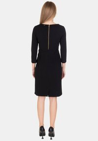 Abito aderente nero con maniche a tre quarti, cerniera posteriore e spacco. Tessuto liscio con linee pulite e dettagli minimi. Scarpe con tacchi alti nere.