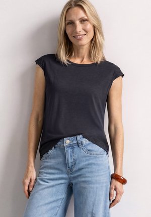 Kvinde med blondt hår iført en sort ærmeløs top, lyseblå jeans og et brunt armbånd, stående mod en ensfarvet lys væg.
