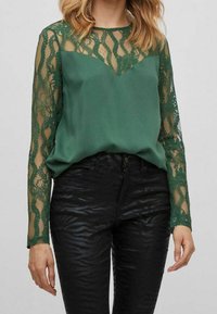Blusa verde con dettagli in pizzo sulle maniche lunghe, caratterizzata da un corpo liscio e una scollatura a V, abbinata a pantaloni con stampa zebra nera.