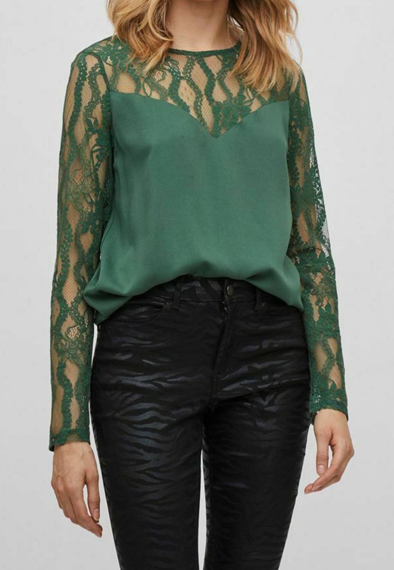Blusa verde con dettagli in pizzo sulle maniche lunghe, caratterizzata da un corpo liscio e una scollatura a V, abbinata a pantaloni con stampa zebra nera.