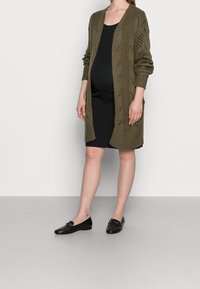Olivově zelený pletený kardigan s otevřeným předním dílem a dlouhými rukávy, navrštěný přes černé bezrukávové šaty a černé mokasíny.