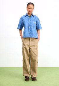 Denim shirt with short sleeves, chest pocket, and button closure. Beige cargo pants with side pockets. Brown shoes. Green flooring background.