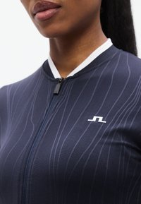 Ναυτικό αθλητικό κορμάκι με διακριτικές κάθετες ρίγες, λευκό γιακά και φερμουάρ στο μπροστινό μέρος. Διαθέτει ένα μικρό λογότυπο σε λευκό στο στήθος.