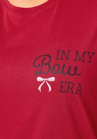 T-shirt rouge avec un texte noir et blanc indiquant "DANS MON ÈRE Bow" accompagné d'un petit graphique de nœud blanc. Fabriqué en tissu doux avec un col rond.