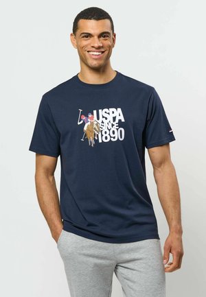 Homme souriant portant un t-shirt bleu marine USPA avec un graphisme de joueur de polo et un pantalon gris, debout avec une main dans la poche.