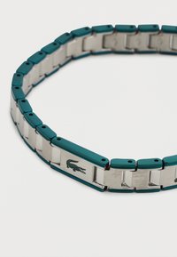 Βραχιόλι από ασημί και τιρκουάζ μεταλλικούς κρίκους με λογότυπο κροκοδείλου σε μία από τις συνδέσεις της αγκράφας, εμφανιζόμενο σε λευκό φόντο.