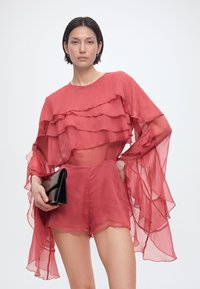 Γυναίκα που φορά διαφανές κόκκινο μπλουζάκι με στρώσεις και φαρδιές μανίκες, συνοδευόμενο από κοντό σορτς σε ταιριαστό χρώμα, κρατώντας μαύρο πατινέ τσαντάκι μπροστά σε απλό φόντο.