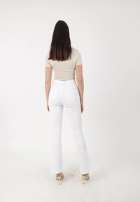 Femme aux longs cheveux bruns portant un haut beige à manches courtes, un jean blanc taille haute évasé et des chaussures à talons beiges, de dos.