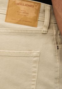 Τσέπη από μπεζ τζιν με δερμάτινο ταμπελάκι ταμπά που γράφει «JACK & JONES Jeans Intelligence Since 1990» στη μέση.