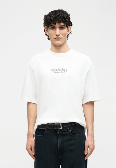 Homme aux cheveux bouclés portant un T-shirt blanc surdimensionné avec un petit dessin au trait noir représentant une table à manger et des chaises, assorti à un jean noir et une ceinture.