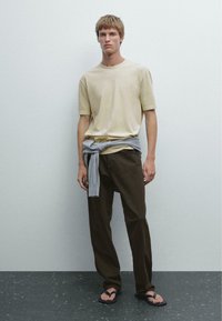 T-shirt di cotone beige, maglione grigio legato in vita, pantaloni marrone scuro e ciabatte nere; su uno sfondo grigio chiaro.