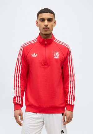 Άνδρας που φορά κόκκινο φούτερ Adidas του Liverpool Football Club με άσπρες ρίγες στα μανίκια και λευκό σορτς, στέκεται μπροστά σε απλό φόντο.