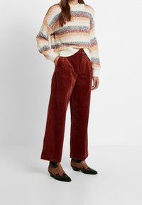 Suéter de punto acogedor y multicolor con rayas horizontales, combinado con pantalones de corduroy de pierna ancha y cintura alta en un profundo tono óxido, y zapatos de punta.