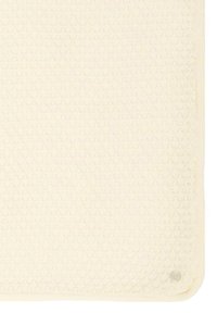 Teksturowana kąpielowa mata w kolorze kremowym z uniesionym wzorem w kształcie komórek pszczelich, otoczona miękką krawędzią z tkaniny. Posiada okrągłą antypoślizgową powłokę w jednym rogu.